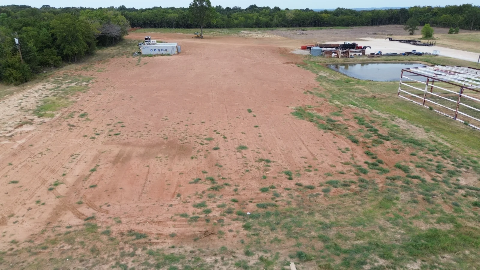 Aerial view of compacted dirt parking lot with metal gates, shipping container, and retention pond