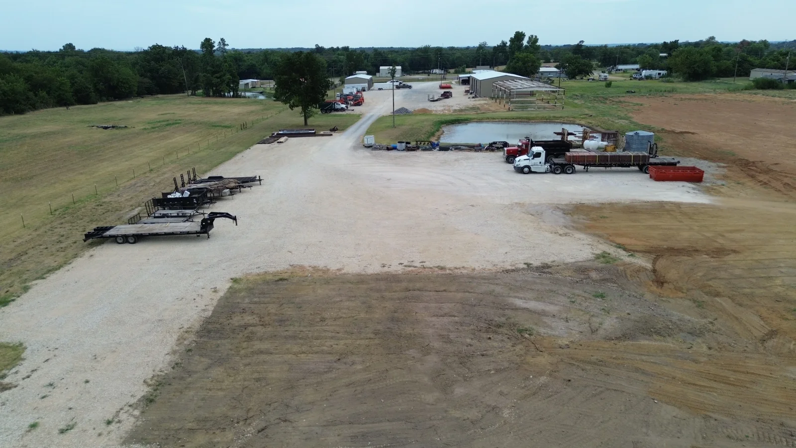 Aerial view of gravel parking lot with equipment trailers, livestock pond, and rural farm buildings